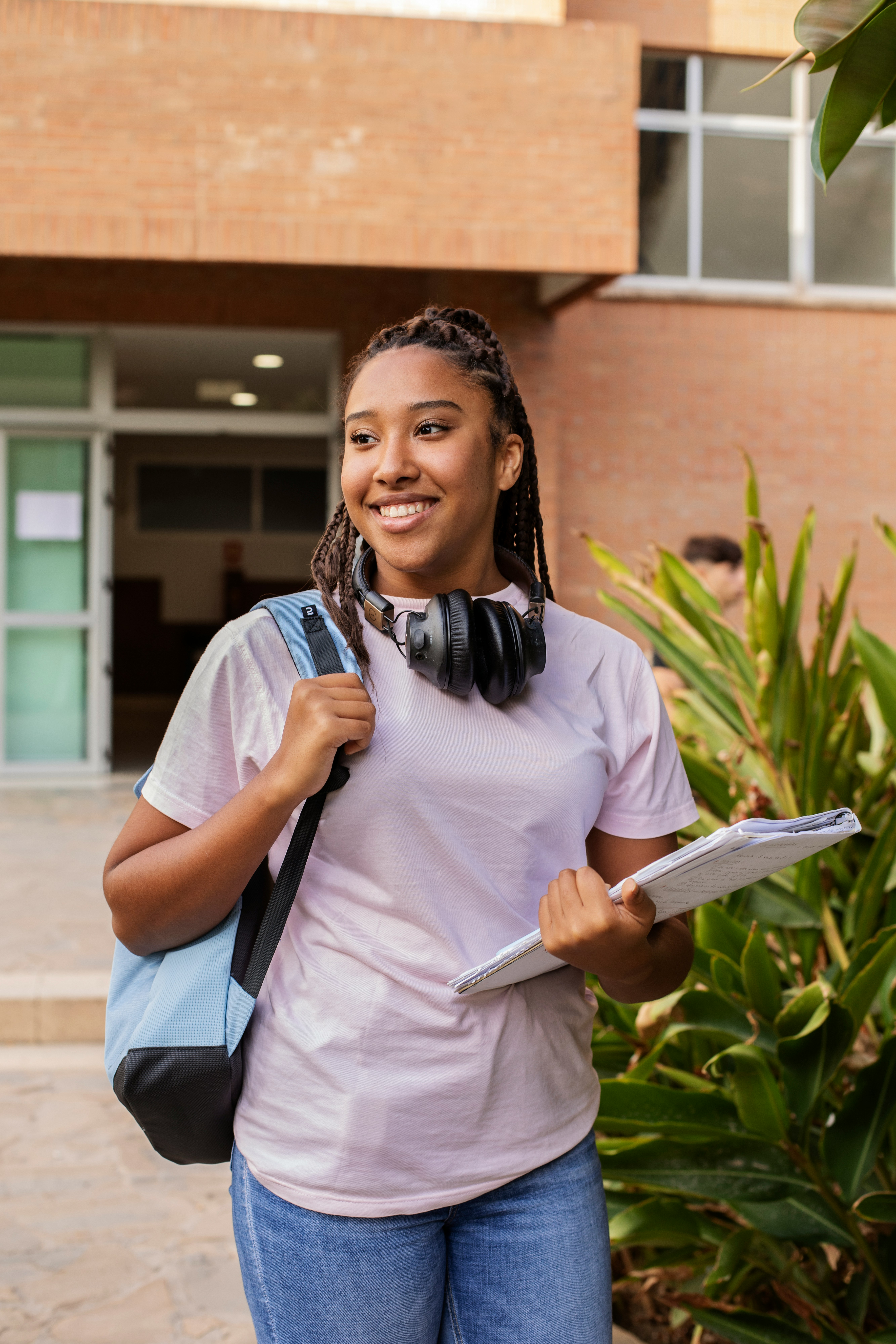 A college student standing outside on campus with a backpack, headphones, and notebooks